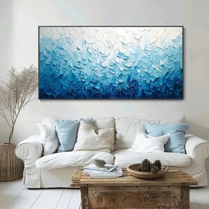 May include: A large abstract painting with a black frame. The artwork features a textured surface with shades of blue and white, creating a gradient effect. The painting is displayed above a white sofa with blue pillows, enhancing the room's decor.