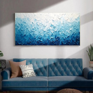 May include: A large rectangular abstract painting with a textured surface. The artwork features a gradient of blue hues, transitioning from white at the top to deep blue at the bottom. The painting is framed and hangs above a blue sofa.