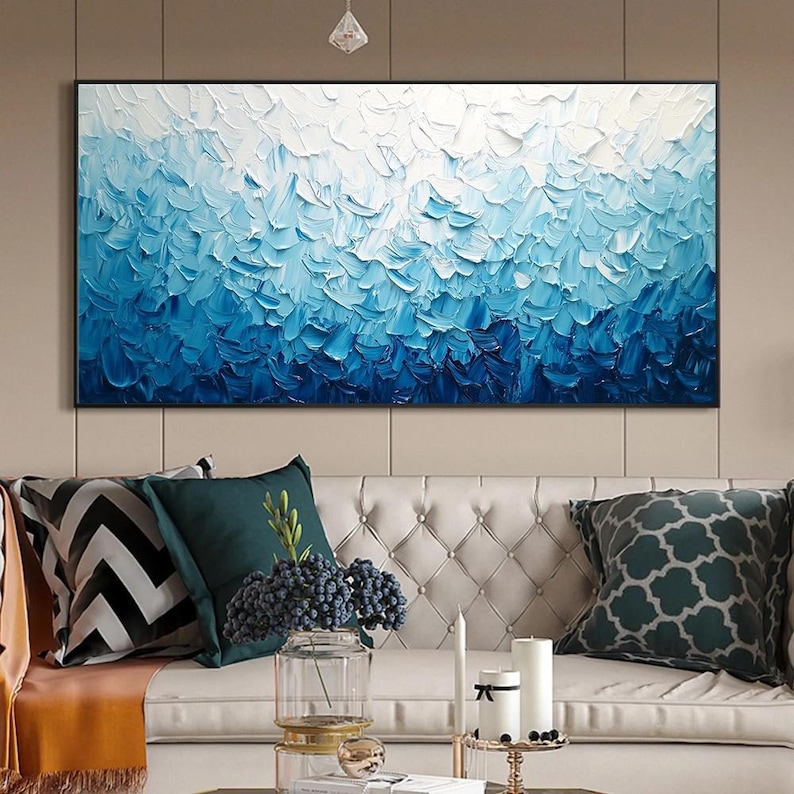 May include: Abstract oil painting in shades of blue and white. The artwork features textured brushstrokes, creating a sense of depth and movement. The painting is framed in a black border and hangs above a white sofa with decorative pillows.