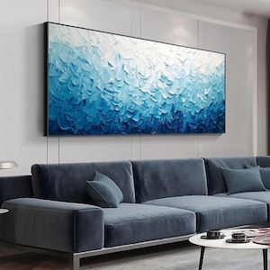 May include: Abstract oil painting in a black frame. The artwork features a textured surface with a gradient of blues, transitioning from white at the top to deep blue at the bottom. The painting hangs above a blue sectional sofa.
