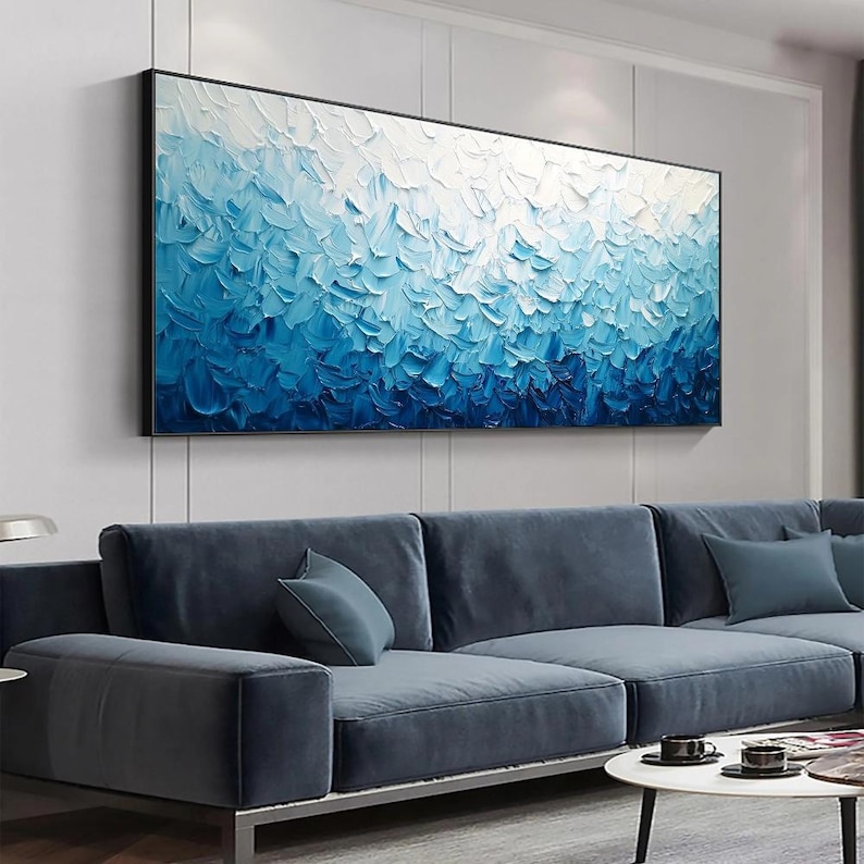 May include: Abstract oil painting in a black frame. The artwork features a textured surface with a gradient of blues, transitioning from white at the top to deep blue at the bottom. The painting hangs above a blue sectional sofa.