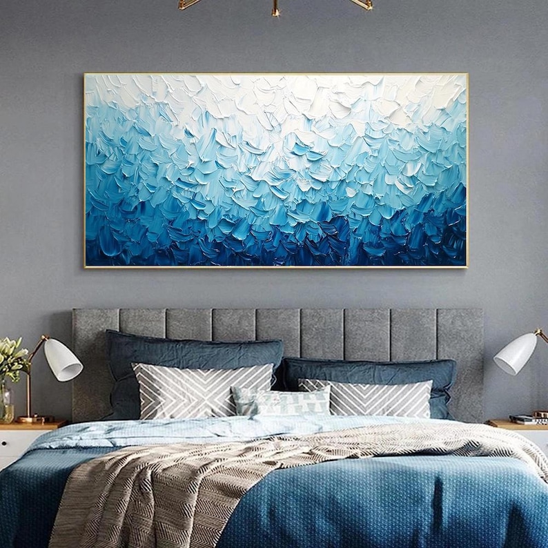 May include: A large, rectangular abstract painting with a gold frame. The artwork features a textured, layered design in shades of blue and white, creating a gradient effect. The painting is displayed above a bed with blue bedding.