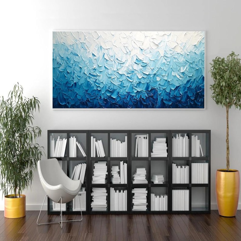 May include: A large, rectangular abstract painting with a white frame. The artwork features a textured, impasto style with shades of blue, transitioning from white at the top to deep blue at the bottom. The painting hangs above a black bookshelf filled with white books.