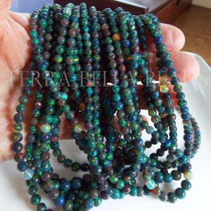 May include: Strands of black opal beads with iridescent flashes of color. The beads are round and appear to be strung together. The image shows multiple strands of the beads, showcasing their dark, shimmering appearance. The text "TERRA BELLA BEADS" is visible in the image.