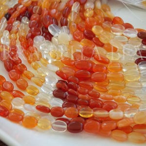 May include: A strand of oval-shaped carnelian beads in various shades of orange, red, and white. The beads are smooth and polished, and they are strung on a white cord.