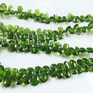 May include: A strand of green gemstone beads, each bead is faceted and shaped like a teardrop. The beads are strung on a clear thread and are arranged in a loose, flowing pattern.