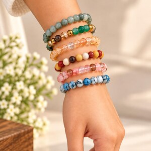 Op de afbeelding: Een stapel van zes kralenarmbanden met diverse edelsteenkleuren, waaronder saliegroen, smaragdgroen, doorschijnend oranje, meerkleurige aardetinten, zachtroze en blauw-witte gespikkelde patronen op een rond elastisch koord.