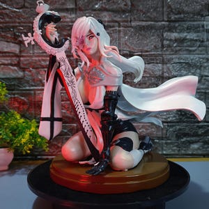 May include: A detailed figurine of a female character with white hair and a flowing white cape. She kneels, holding a large sword with intricate designs. The figure is on a brown base, with a brick wall backdrop.