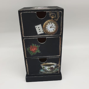 Könnte beinhalten: Eine schwarze Aufbewahrungskommode aus Holz mit drei Schubladen. Die oberste Schublade zeigt eine goldene Taschenuhr und eine Spielkarte. Die mittlere Schublade ist mit einer roten Rose verziert. Die untere Schublade zeigt eine Teetasse.