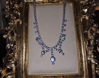 May include: A light blue rhinestone necklace displayed on a gold ornate picture frame. The necklace features a series of small, round rhinestones connected by delicate links, with larger stones at the focal points. The frame has a detailed, vintage design.