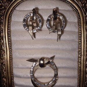 May include: A vintage jewelry set featuring a pair of earrings and a brooch. The earrings and brooch have a gold-tone metal finish with clear, rectangular-cut stones set in a circular design. The earrings have a unique, abstract shape.