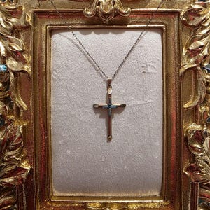 May include: A gold-toned cross pendant with a delicate chain, displayed within a decorative gold frame. The cross features a detailed design with what appears to be blue and white accents. The frame has ornate carvings.