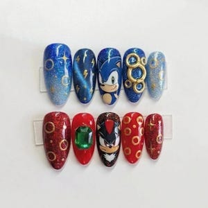 May include: A set of ten press-on nails featuring blue and red glitter finishes. The blue nails display Sonic the Hedgehog, gold rings, and star motifs. The red nails feature Shadow the Hedgehog, a large green gemstone, and gold ring patterns.