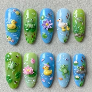 May include: A set of ten artificial nails with a water-themed design. Each nail features a different miniature pond scene with elements like ducks, frogs, fish, lily pads, and dragonflies. The nails are painted in shades of blue and green.