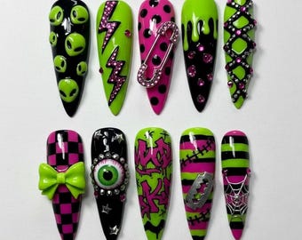 May include: A set of ten stiletto-shaped press-on nails with various designs. The nails feature black, neon green, and pink colors with embellishments like aliens, lightning bolts, polka dots, a safety pin, dripping paint, bows, an eyeball, graffiti, a razor blade, and a spiderweb.