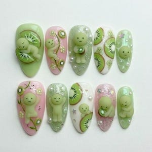 May include: A set of ten almond-shaped artificial nails with a kiwi fruit and baby-like figure design. The nails feature a variety of colours, including green, pink, and white, with pearl and star accents. The kiwi fruit slices and figures are 3D.