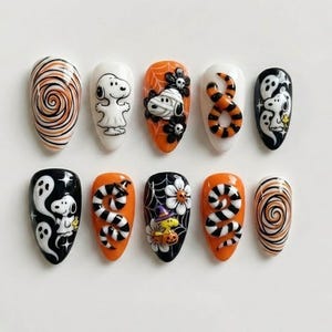 May include: A set of ten press-on nails featuring Halloween-themed designs including Snoopy, Woodstock, ghosts, orange and black swirls, striped snakes, spider webs, flowers, and skulls in a vibrant palette of orange, black, white, and yellow.