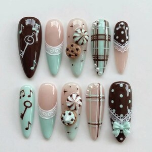 Op de afbeelding: Een set van tien amandelvormige kunstnagels in een kleurenpalet van bruin, mintgroen en nude. De ontwerpen bevatten muzieknoten, sleutels, witte kantpatronen, ruitjes, kleine parels, 3D-koekje en glazuur bedels en een mintgroene strik.