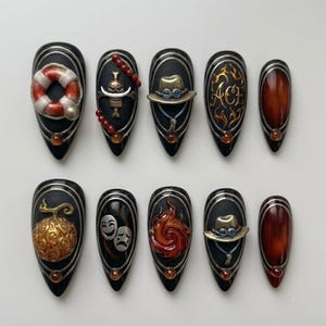 May include: A set of ten black almond-shaped press-on nails featuring 3D embellishments including a lifebuoy, a skull emblem, a hat, the text ACE, a golden fruit, comedy and tragedy masks, and a swirling flame, all accented with silver lines and gems.