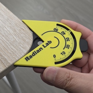 May include: A yellow Radian Lab angle finder tool is held against the corner of a light wood surface. The tool has a black dial with degree markings from 0 to 50. The text "Radian Lab" is printed on the tool.