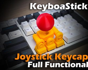 May include: A yellow and red joystick keycap, labeled "KeyboaStick" and "Joystick Keycap Full Functional", is mounted on a white and black keyboard. The joystick has a red ball on top and yellow supports.