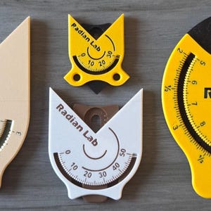 May include: A collection of Radian Lab angle finders in various sizes and colors. The tools are beige, white, and yellow, with black and green accents. Each tool features a curved scale with measurements in inches and centimeters. The text "Radian Lab" is printed on each tool.