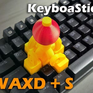 May include: A custom keyboard with black keys and a yellow and red decorative figure on top. The text "KeyboaStick" is at the top, and "WAXD + S" is at the bottom in orange.