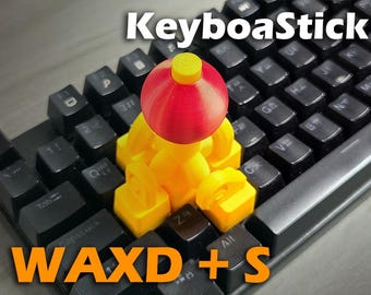 May include: A custom keyboard with black keys and a yellow and red decorative figure on top. The text "KeyboaStick" is at the top, and "WAXD + S" is at the bottom in orange.