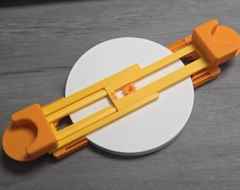 May include: An adjustable circle cutter in yellow and orange, with a white circular base. The tool features a gear mechanism and is designed for cutting circles. It's made of plastic and suitable for crafting or DIY projects. The white circle is approximately 10 cm in diameter.