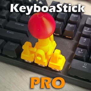 May include: A yellow and red KeyboaStick PRO device is mounted on a black keyboard. The device has a red sphere on top and yellow supports. The word "KeyboaStick" is at the top, and "PRO" is at the bottom.