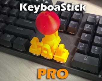 May include: A yellow and red KeyboaStick PRO device is mounted on a black keyboard. The device has a red sphere on top and yellow supports. The word "KeyboaStick" is at the top, and "PRO" is at the bottom.