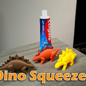 May include: Three dinosaur-shaped toothpaste tube squeezers in brown, orange, and yellow. The orange dinosaur is positioned holding a blue and red Colgate toothpaste tube. Each dinosaur features a spiky back and four legs.