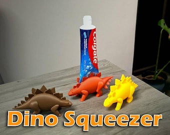 May include: Three dinosaur-shaped toothpaste tube squeezers in brown, orange, and yellow. The orange dinosaur is positioned holding a blue and red Colgate toothpaste tube. Each dinosaur features a spiky back and four legs.