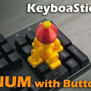 Puede incluir: Un accesorio KeyboaStick NUM with Button amarillo y rojo está conectado a un teclado negro. El accesorio tiene una parte superior roja y una base amarilla. Las teclas del teclado son negras con letras blancas. El texto "KeyboaStick" y "NUM with Button" son visibles.