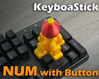 May include: A yellow and red KeyboaStick NUM with Button accessory is attached to a black keyboard. The accessory has a red top and yellow base. The keyboard keys are black with white lettering. The text "KeyboaStick" and "NUM with Button" are visible.