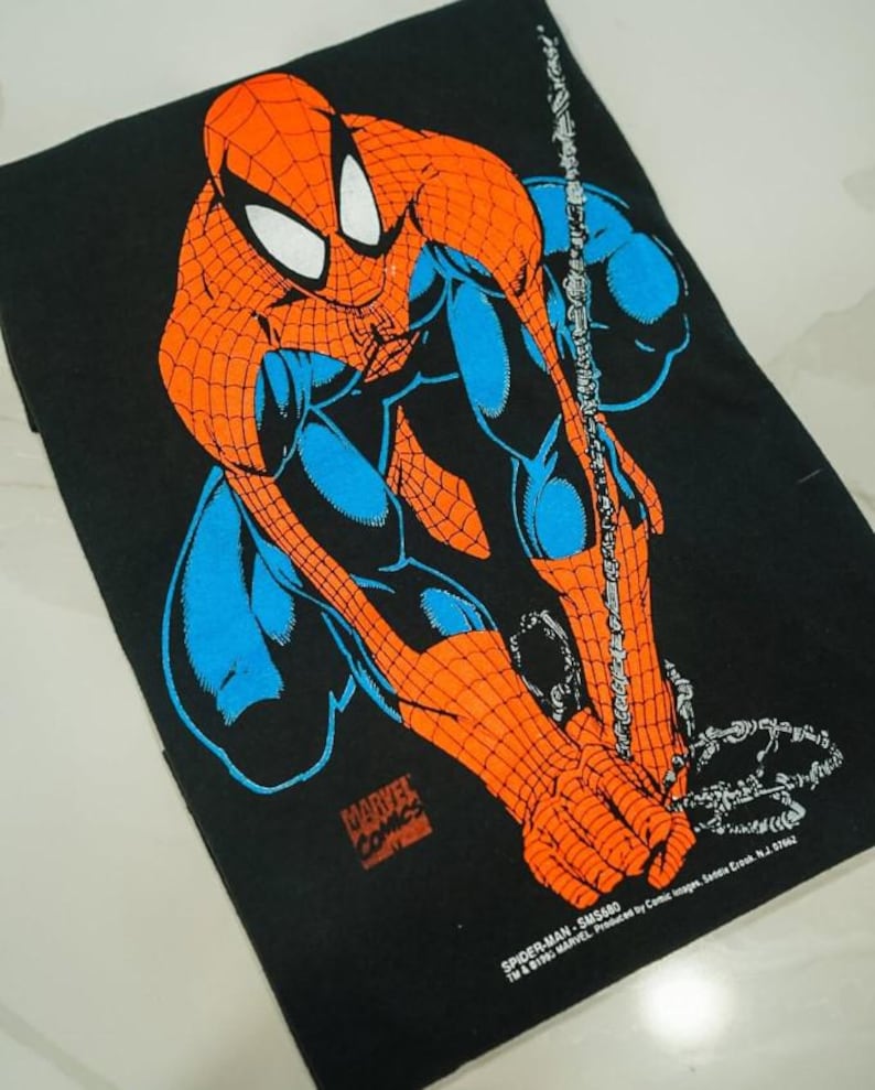 May include: A black t-shirt featuring a graphic print of Spider-Man in a crouching pose with orange and blue colors. The print includes the text Marvel Comics, Spider-Man, SMS680, TM & &copy;1991 MARVEL Produced by Comic Images, Saddle Brook, N.J. 07662.