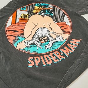 May include: A charcoal gray t-shirt featuring a circular graphic on the back. The illustration depicts a comic book character with long white hair and a black mask, kneeling on a bed with orange bedding and a teal blanket. The text SPIDER-MAN is printed below.