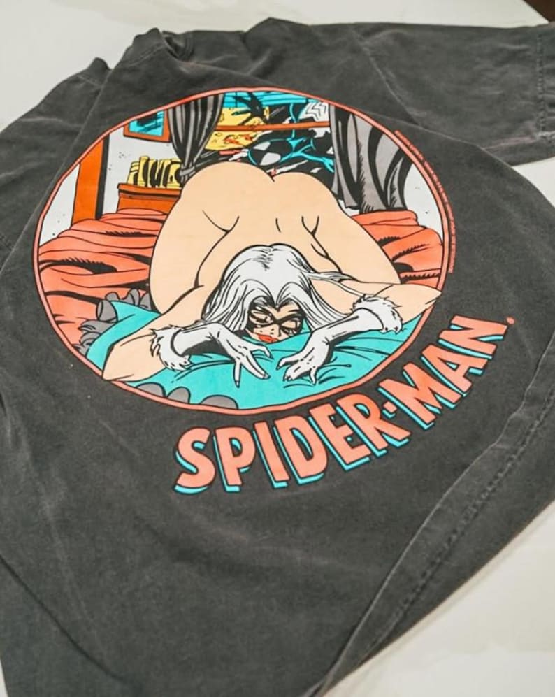 May include: A charcoal gray t-shirt featuring a circular graphic on the back. The illustration depicts a comic book character with long white hair and a black mask, kneeling on a bed with orange bedding and a teal blanket. The text SPIDER-MAN is printed below.