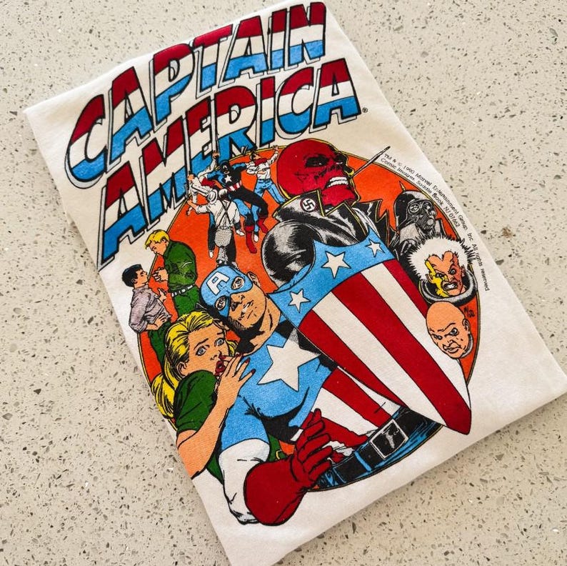 May include: A white t-shirt featuring a vintage Captain America comic book graphic. The print displays Captain America with his shield, a blonde character, and various villains including Red Skull. The text reads CAPTAIN AMERICA at the top.