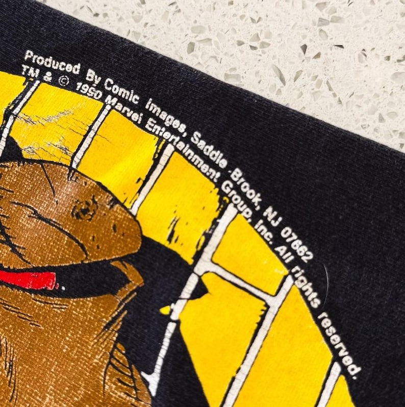 K&ouml;nnte beinhalten: Eine schwarze Stoffoberfl&auml;che mit einer gelben kreisf&ouml;rmigen Grafik, wei&szlig;en Gitterlinien und einer braunen Charakterillustration. Der Text lautet: Produced By Comic Images, Saddle Brook, NJ 07662 TM & &copy; 1990 Marvel Entertainment Group, Inc. All rights reserved.