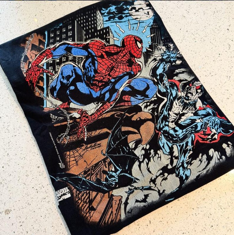 May include: A black t-shirt featuring a vibrant comic book graphic of Spider-Man crouching on a rooftop above a city skyline at night. The illustration includes a dark creature with wings, a full moon, and the text Marvel Comics and Spider-Man.