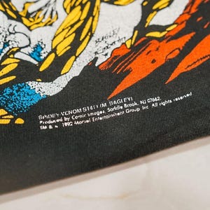 May include: A black t-shirt featuring a graphic illustration with yellow, orange, and blue colors. The print includes the text: SPIDEY VENOM S1411 (M BAGLEY) Produced by Comic Images, Saddle Brook, NJ 07662. TM & 1992 Marvel Entertainment Group Inc.