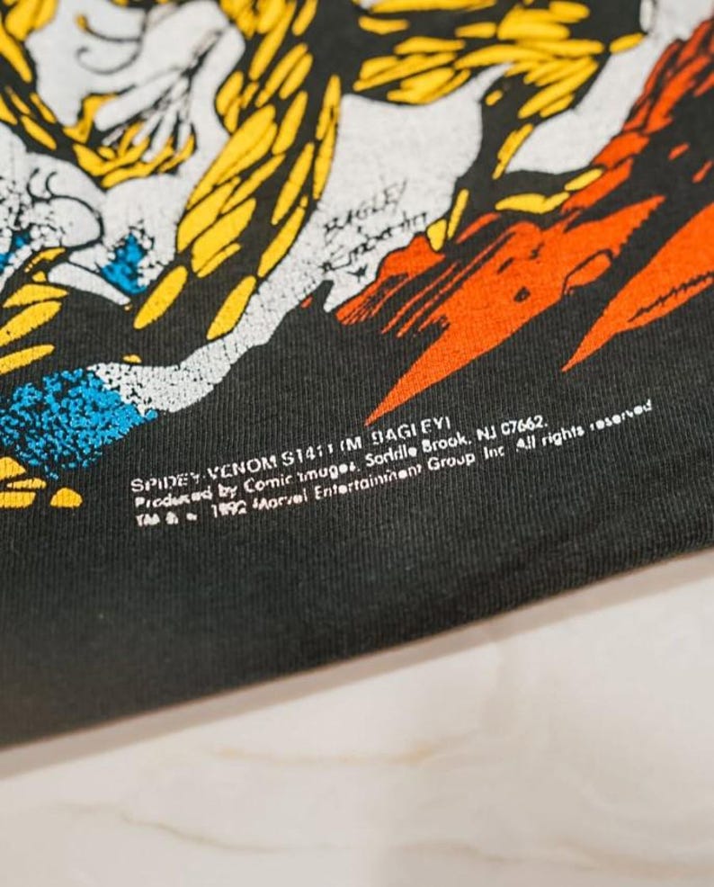May include: A black t-shirt featuring a graphic illustration with yellow, orange, and blue colors. The print includes the text: SPIDEY VENOM S1411 (M BAGLEY) Produced by Comic Images, Saddle Brook, NJ 07662. TM & 1992 Marvel Entertainment Group Inc.