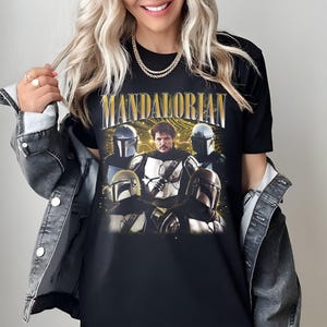 K&ouml;nnte beinhalten: Schwarzes T-Shirt mit einer Grafik von Charakteren aus der Mandalorian-Serie. Das Wort "MANDALORIAN" ist oben in goldfarbenen Buchstaben aufgedruckt. Das Design enth&auml;lt Bilder des Mandalorianers und anderer Charaktere mit Helmen.