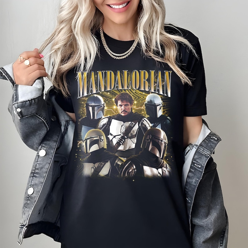 K&ouml;nnte beinhalten: Schwarzes T-Shirt mit einer Grafik von Charakteren aus der Mandalorian-Serie. Das Wort "MANDALORIAN" ist oben in goldfarbenen Buchstaben aufgedruckt. Das Design enth&auml;lt Bilder des Mandalorianers und anderer Charaktere mit Helmen.