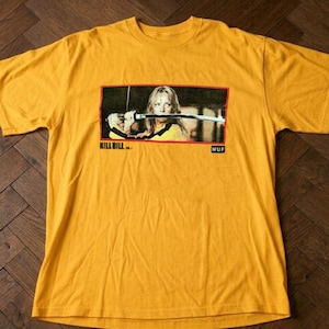 Op de afbeelding: Mosterdgeel t-shirt met een rechthoekige afbeelding uit de film Kill Bill. De afbeelding toont een vrouw die een zwaard vasthoudt. De tekst "KILL BILL" en "HUF" zijn op het t-shirt gedrukt.