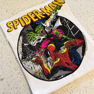 May include: A white t-shirt featuring a circular graphic of Spider-Man fighting a green lizard-like creature. The text SPIDER-MAN is printed in large yellow letters with a red outline above the circular illustration, which includes black and white brick details.