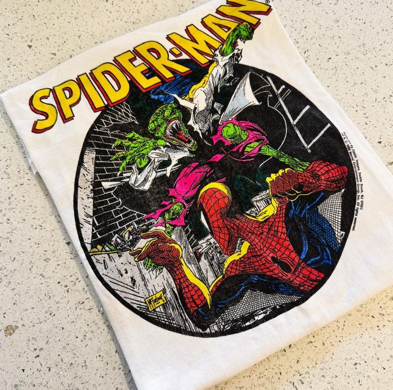 May include: A white t-shirt featuring a circular graphic of Spider-Man fighting a green lizard-like creature. The text SPIDER-MAN is printed in large yellow letters with a red outline above the circular illustration, which includes black and white brick details.