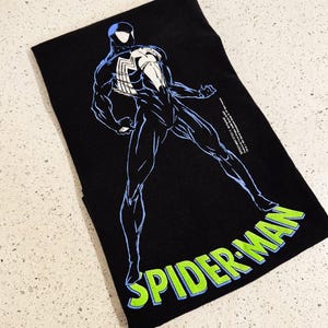 May include: A black t-shirt featuring a graphic of a character in a black suit with a large white spider emblem on the chest. The character is outlined in light blue. The text SPIDER-MAN is printed in bright green with a light blue outline below.