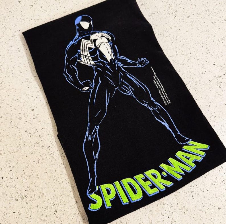 May include: A black t-shirt featuring a graphic of a character in a black suit with a large white spider emblem on the chest. The character is outlined in light blue. The text SPIDER-MAN is printed in bright green with a light blue outline below.
